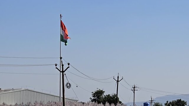 A tricolor Indian flag flies proudly above a network of utility poles power lines, manufacturing industry building. The flag's colors represent strength, peace, and growth, embodying national signific