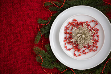 Elegant holiday table setting featuring white plates adorned with golden floral decoration and red beads, surrounded by fresh evergreen branches on a vibrant red background