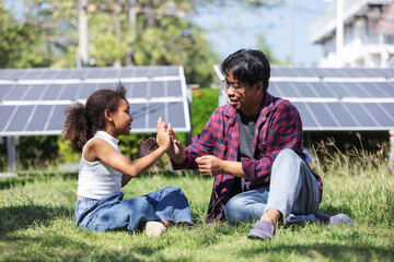 A father and daughter sit on green grass near solar panels, sharing a joyful high five while learning about clean energy.