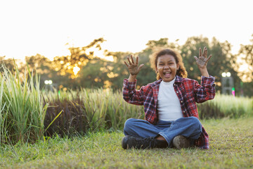 A joyful young child sits on grass near a rice field with muddy hands raised, laughing freely.