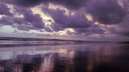 Fototapeta premium ​A moody coastal scene featuring heavy dark storm clouds over a reflective beach, with crashing white waves under dim light