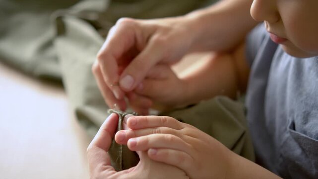 Mother repairing hem with needle while toddler stays close wanting to help. Everyday family moment, sewing concept, patience, handmade craftwork, and care in home environment.