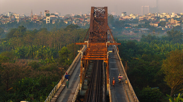 Long Bien Bridge at sunrise with a large glowing red sun rising above the historic 100-year-old iron structure in the early morning light of Hanoi.