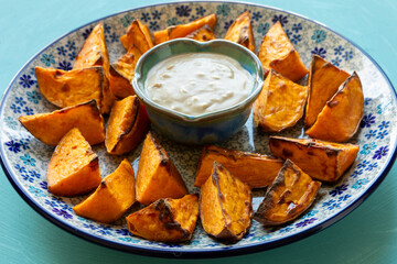 Baked sweet potatoes on a platter, with tahini dip in a small bowl in the middle. Blue background.