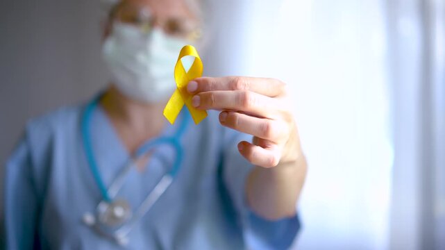 A female doctor in medical uniform holds a yellow satin ribbon, symbol for suicide prevention, bone cancer, endometriosis, and childhood cancer awareness.
