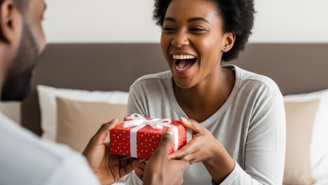 Smiling woman receiving red gift box from man. Happy african american couple celebrating anniversary at home. Birthday surprise present concept. - Powered by Adobe