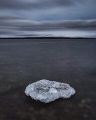 At lake Femunden in Norway, a piece of ice floats on the water surface. The sky is cloudy and grey, reflecting the cool atmosphere of early morning. Mountains are visible in the background.