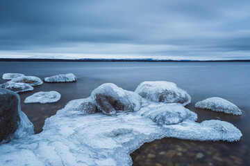Ice forms at the shoreline of lake Femunden in Norway during winter season with rocks visible above the frozen surface