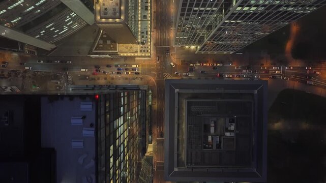 Aerial top down view of a financial district at night, flying between modern skyscrapers with illuminated windows and foggy streets with traffic