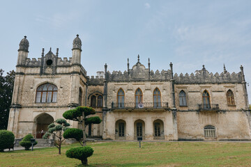 Fototapeta premium Majestic historic castle with ornate stone facade and tall arches rises over a manicured lawn with trimmed trees under a clear sky, evoking timeless architectural grandeur.