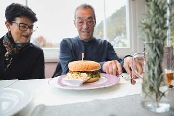 Senior couple dining together at a restaurant, sharing a large gourmet burger with coleslaw, smiling and celebrating a relaxed lunch date and happy, authentic moment together