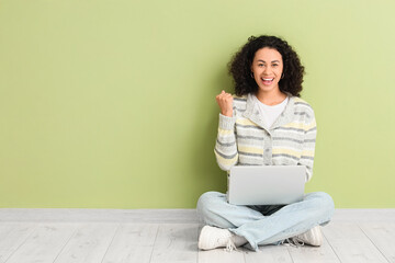 Happy young African-American woman working with laptop near green wall
