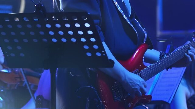 Musician plays red electric guitar during performance. Music stand and sheet music indicate structured musical setting with blue light