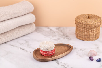 Two natural solid shampoo bars on a wooden tray with beige towels, wicker basket and mineral stones. Peach background and marble table. Horizontal image with copy space. 