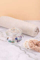 Front view vertical composition of glass jar with colorful decorative stones next to beige towel and textile scrunchie on marble surface, peach background. Concept of natural self care and wellness