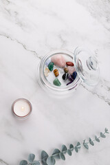 Top view vertical of glass jar with colorful natural stones, lit candle and eucalyptus branch on white marble background. Copy space. Concept of holistic wellness and self care