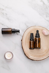 Vertical flat lay of amber glass cosmetic bottles on wooden base with lit candle and rose quartz, on light marble background. Copy space. Concept of natural self care and wellness