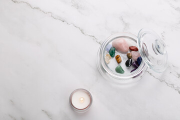 Top view horizontal of glass jar with natural stones and lit candle on light marble background. Minimal and calming style with ample copy space. Concept of wellness, balance and self care