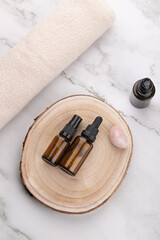 Vertical flat lay of amber cosmetic bottles on wooden base with beige towel and rose quartz stone on light marble background. Copy space. Concept of natural self care and wellness