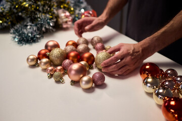Hands arranging Christmas New Year baubles on white table with tinsel and warm bokeh lights, metallic red gold pink palette, careful styling by home decorator, festive craft mood