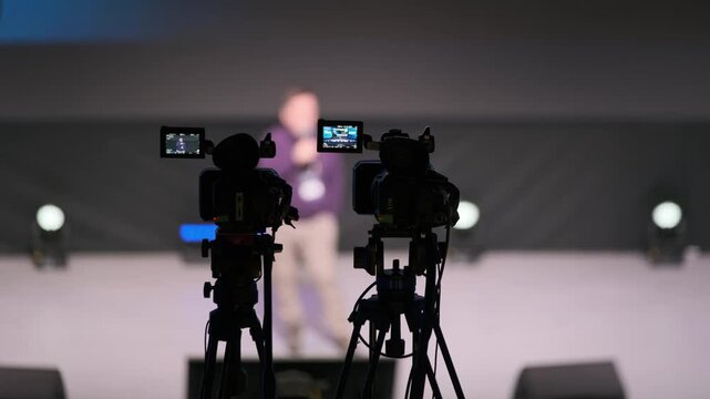 Cameras on tripods capture a blurred speaker on stage during a live event with stage lights and dark backdrop