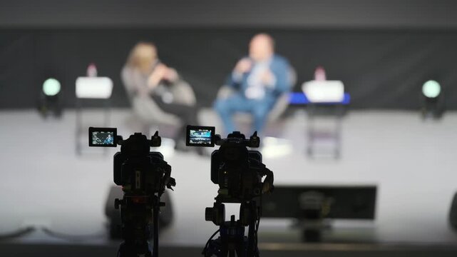 Two blurred speakers on stage answer questions during talk while two cameras capture scene from close up creating professional panel impression