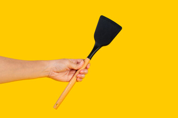 Hand with Black plastic kitchen spatula, kitchenware for cooking on yellow background.