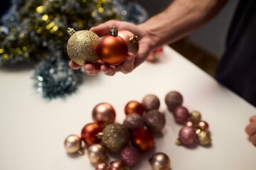 Hands arranging Christmas New Year baubles on white table with tinsel and warm bokeh lights, metallic red gold pink palette, careful styling by home decorator, festive craft mood