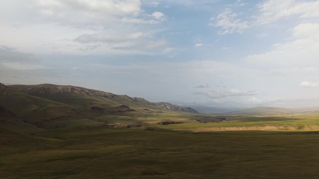 Kyrgyzstan - Soft Sunlight Sweeps Across Wide Green Plains Framed by Distant, Rugged Hills Under a Calm Sky - Aerial Drone Shot