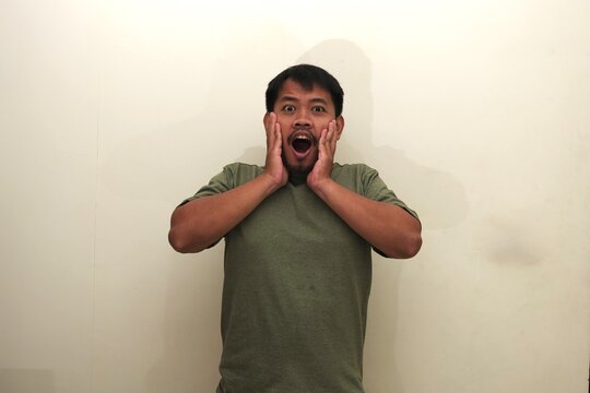 Portrait of a Southeast Asian man showing extreme surprise or shock, hands on cheeks and mouth open, isolated, ideal for emotion or ad use.

