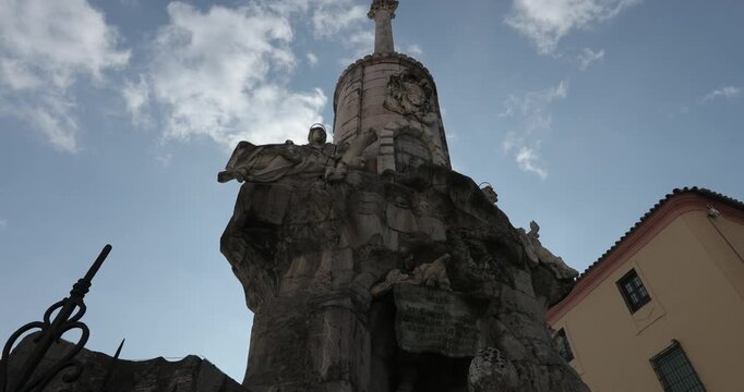 Dramatic low-angle shot of an ornate, historic stone monument or pillar with statues and carvings, standing against a bright blue sky with patchy white clouds in a European city square