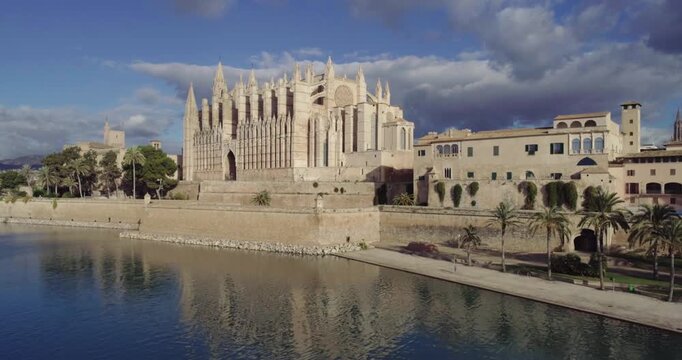 Majestic low aerial view of Palma Cathedral (La Seu) in Mallorca, Spain. The shot highlights the Gothic cathedral's architecture, seaside fortress walls, and surrounding historic city buildings under 