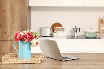 Vase with artificial flowers and laptop on table in kitchen