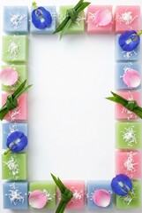 Thai dessert Khanom Chan flat lay frame of pastel layer cakes with butterfly pea flowers and pandan leaves on vertical white background with coconut topping and copy space for menu design.