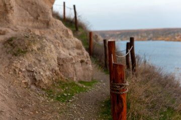 Rustic wooden fence lining a narrow path along a rocky cliff, overlooking a tranquil river under soft daylight.