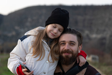 Smiling bearded man and girl in warm clothes, enjoying a tender moment together outside with blurred natural background.
