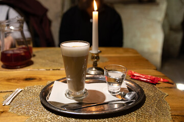 Glass of latte and water served on a silver tray beside a lit candle, creating a warm and cozy atmosphere on a rustic wooden table.