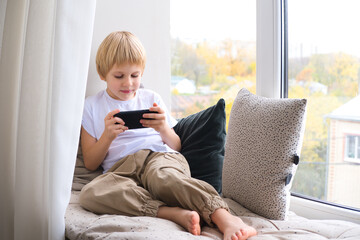 blonde little boy using smartphone sitting on the windowsill
