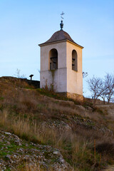 Historic stone bell tower stands on a grassy hillside under a clear blue sky, surrounded by bare trees and rustic scenery.