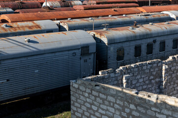 A row of weathered train cars with rusted roofs sits behind a stone wall in bright sunlight, evoking themes of decay, history, and industrial heritage.