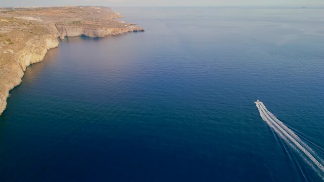 Aerial 4K tracking shot of a boat cruising along the majestic white Dingli Cliffs, white water trail in turquoise Mediterranean sea, scenic Malta coastline, drone
