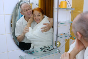 Happy Senior Couple Making Daily Beauty Routine In Bathroom At Home, Smiling Mature Spouses Embracing And Looking In Mirror, Getting Ready Together In The Morning, Enjoying Selfcare.