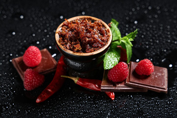 Hookah bowl of tobacco with berries, chocolate pieces, dried chili peppers and mint leaves on black background