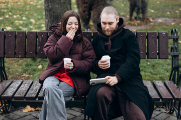 Young couple sitting on park bench drinking coffee and using smartphone.