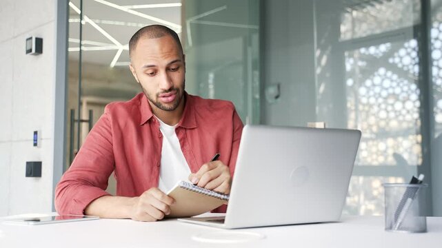 Businessman watching video call conference using a laptop computer takes notes while sitting at workplace in a modern business office. Confident manager communicates remotely at an online meeting