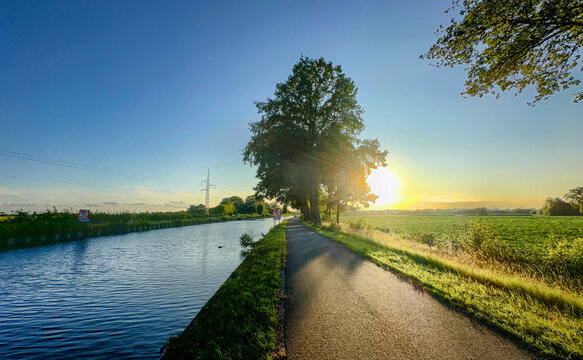 Golden hour canal path bathed in warm sunlight, long tree shadows stretching across lane, peaceful route suggesting cycling