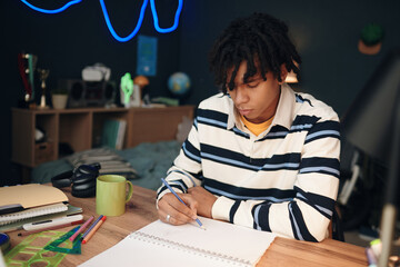 Black teenage boy sitting at desk writing in notebook with focused expression, surrounded by school supplies in bedroom, studying or doing homework for academic assignment