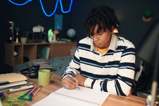 Black teenage boy sitting at desk writing in notebook with focused expression, surrounded by school supplies in bedroom, studying or doing homework for academic assignment