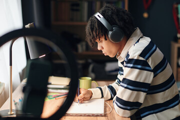 Black teenager boy wearing headphones sitting at desk studying and writing in notebook, focused on learning with tablet and school supplies nearby, bookshelf in background