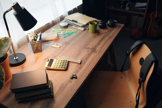 Wooden desk holding calculator, books, stationery, green mug, headphones, and plant near window, empty chair with backpack hanging on backrest, lamp light illuminating workspace